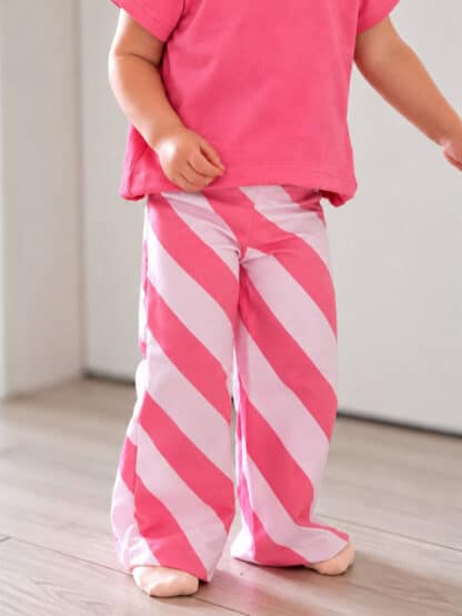 Rosa-weiß schräg gestreifte Wide-Leg-Hose mit pinkem T-Shirt, barfuß auf dem Holzboden.