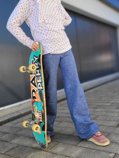 Blaue weite Jogginghose in Jeansoptik, dazu ein Skateboard in der Hand auf dem Gehweg.