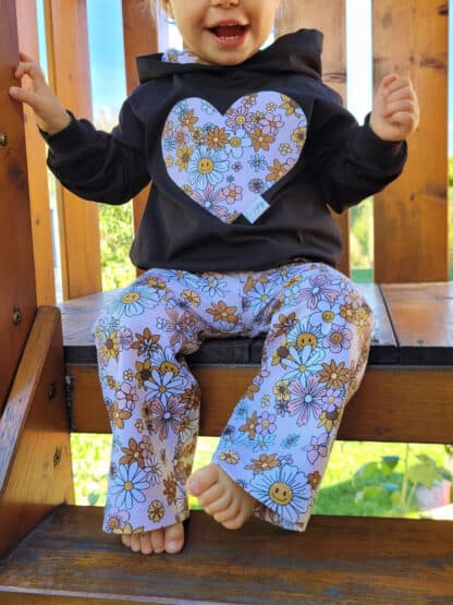 Sitzend im Spielhaus: selbst genähte weite Hose mit bunten Blumen und Herz-Pullover.