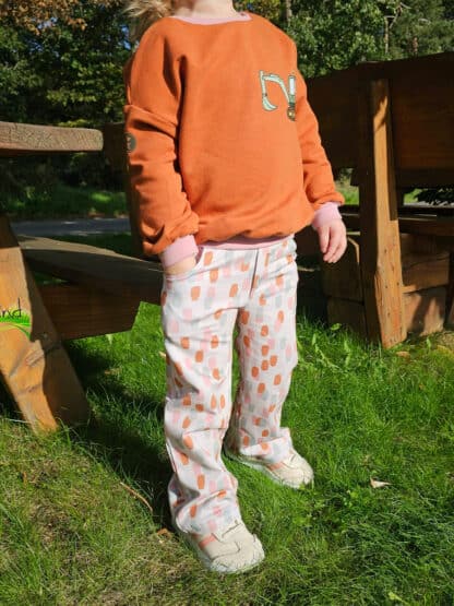 Selbst genähte weite Hose mit orangem Muster und orangem Pullover vor einer Holzbank.