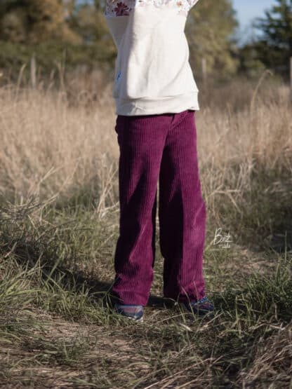 Weinrote Wide-Leg-Hose aus Cordjersey mit weißem Pullover im hohen, trockenen Gras.
