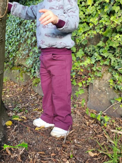 Beerenfarbene Wide-Leg-Hose mit grauem Pullover vor einer efeubewachsenen Mauer.