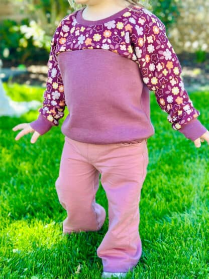 Rosa selbst genähte Wide-Leg-Hose am Kleinkind mit geblümtem Pullover auf dem Rasen.
